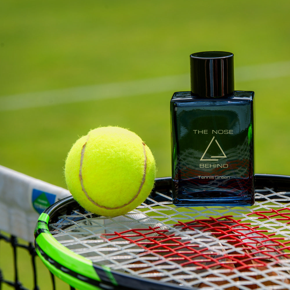 Parfumflakon „Tennis Green“ von „The Nose Behind“ steht auf einem Tennisschläger neben einem gelben Tennisball.