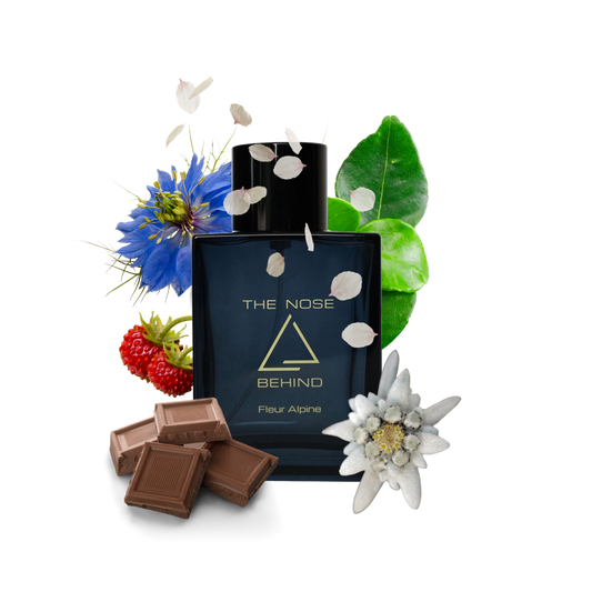 Parfümflakon „Fleur Alpine“ von „The Nose Behind“ umgeben von natürlichen Duftnoten: blauer Blüte, Edelweiß, grünen Blättern, Erdbeere, Schokoladenstücken und herabfallenden Blütenblättern. Die Bildkomposition symbolisiert die Duftkomponenten des Parfüms – floral, fruchtig und gourmand.